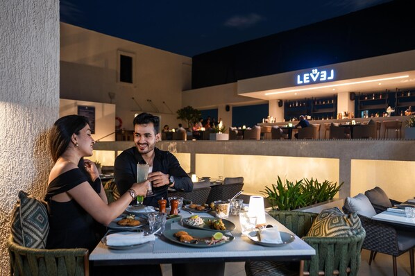 Un área de comedor al aire libre con mesas y sillas, un bar al fondo y un cartel que dice "LEVEL".