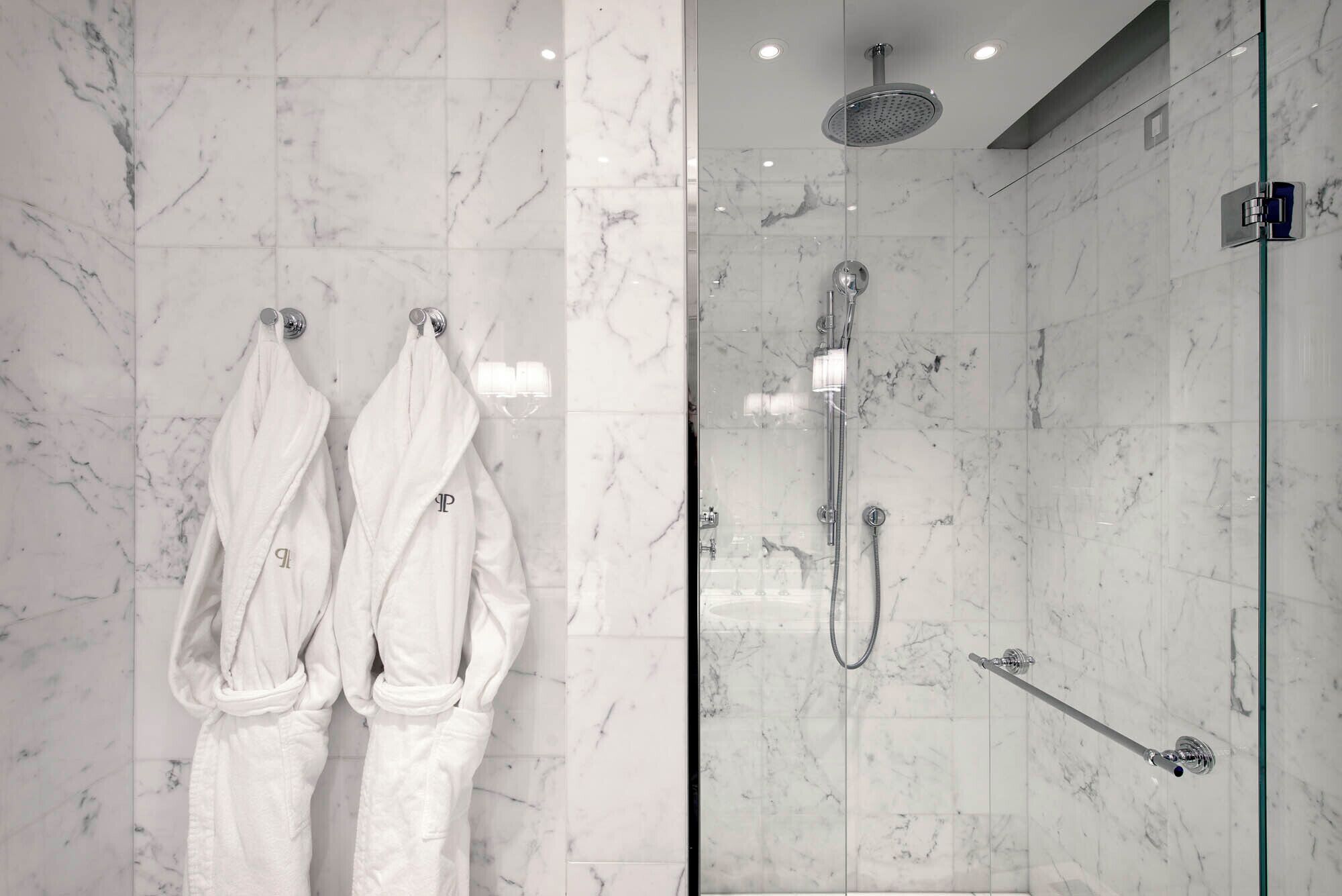 Une salle de bain aux murs recouverts de marbre, avec une douche en verre et deux peignoirs blancs suspendus à des crochets.