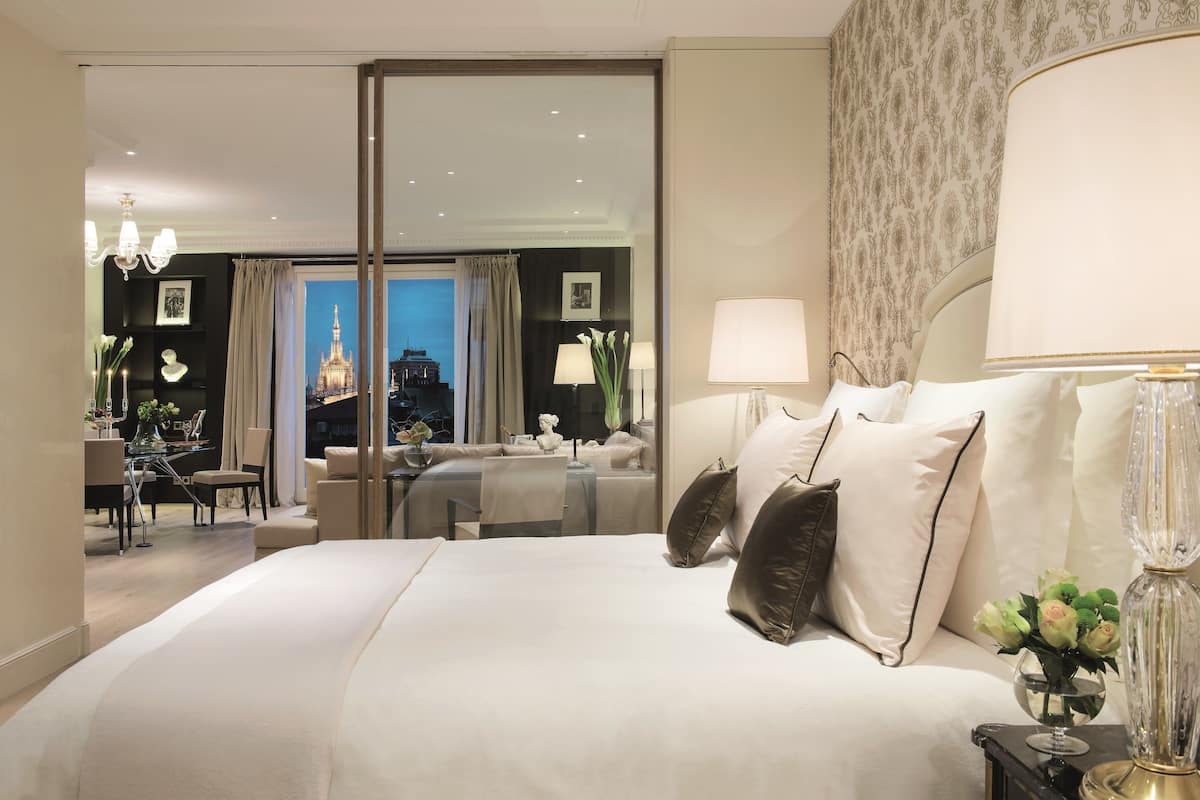 Ein Hotelzimmer mit einem großen Bett, einem Schreibtisch mit Stuhl, einer Couch und Blick auf eine Stadtlandschaft durch eine Schiebetür.