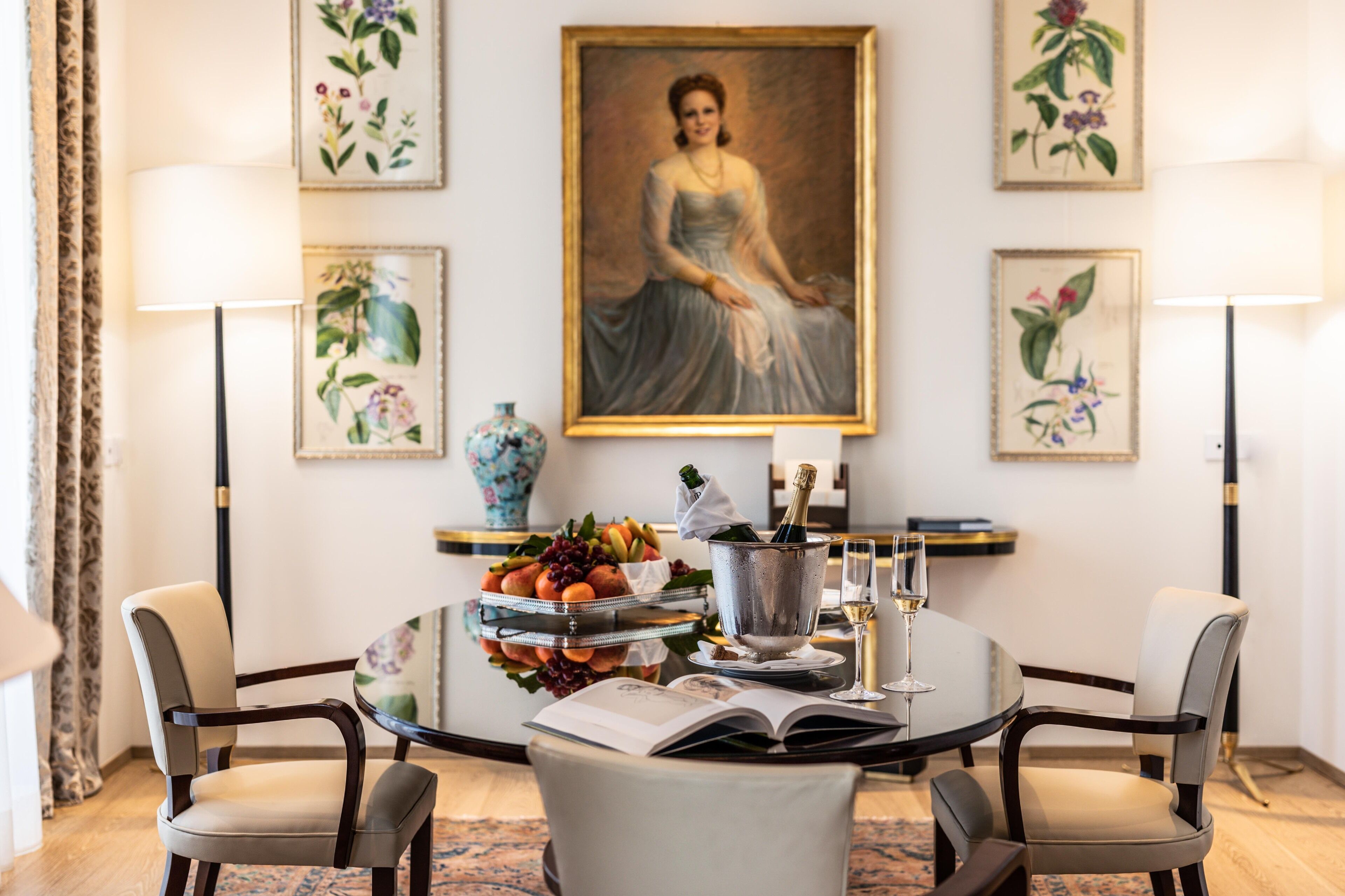 Een eetgedeelte met een ronde tafel, twee stoelen, een vaas met bloemen, een fles champagne en een boek.