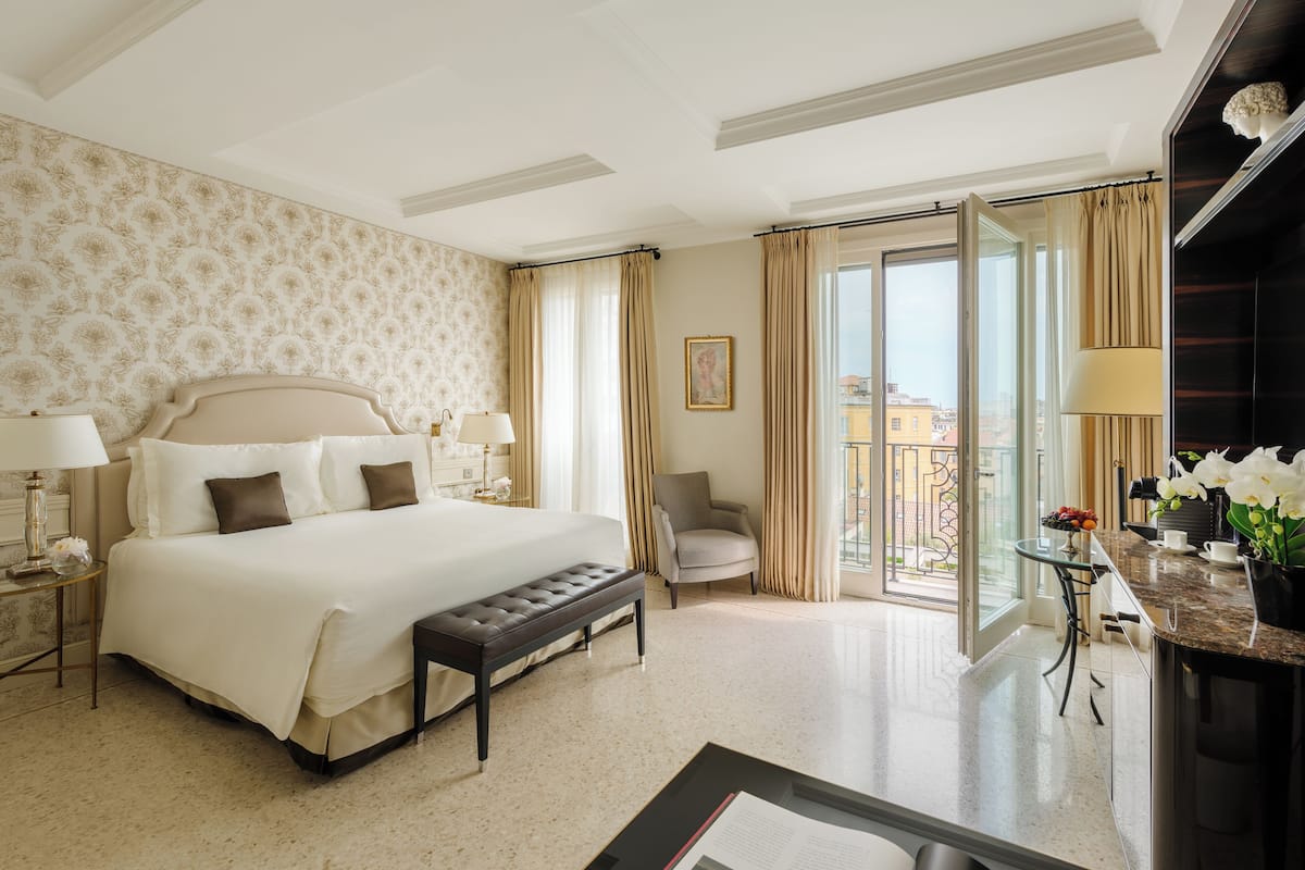 Ein Hotelzimmer mit einem großen Bett, einer Bank, einem Sessel, einem Schreibtisch mit Lampe und Blick auf die Stadt durch das Fenster.