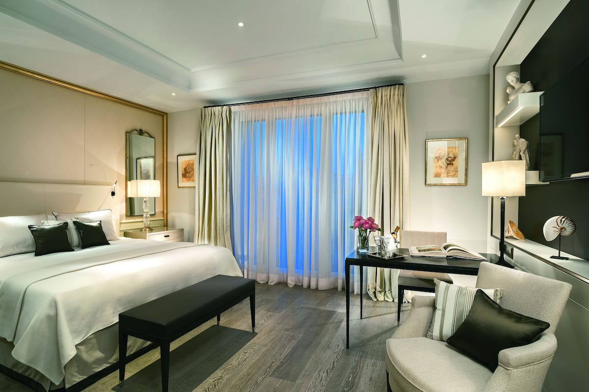 Ein Hotelzimmer mit einem großen Bett, einem Schreibtisch, einem Sessel, einer Bank und einem Fenster mit transparenten Vorhängen.