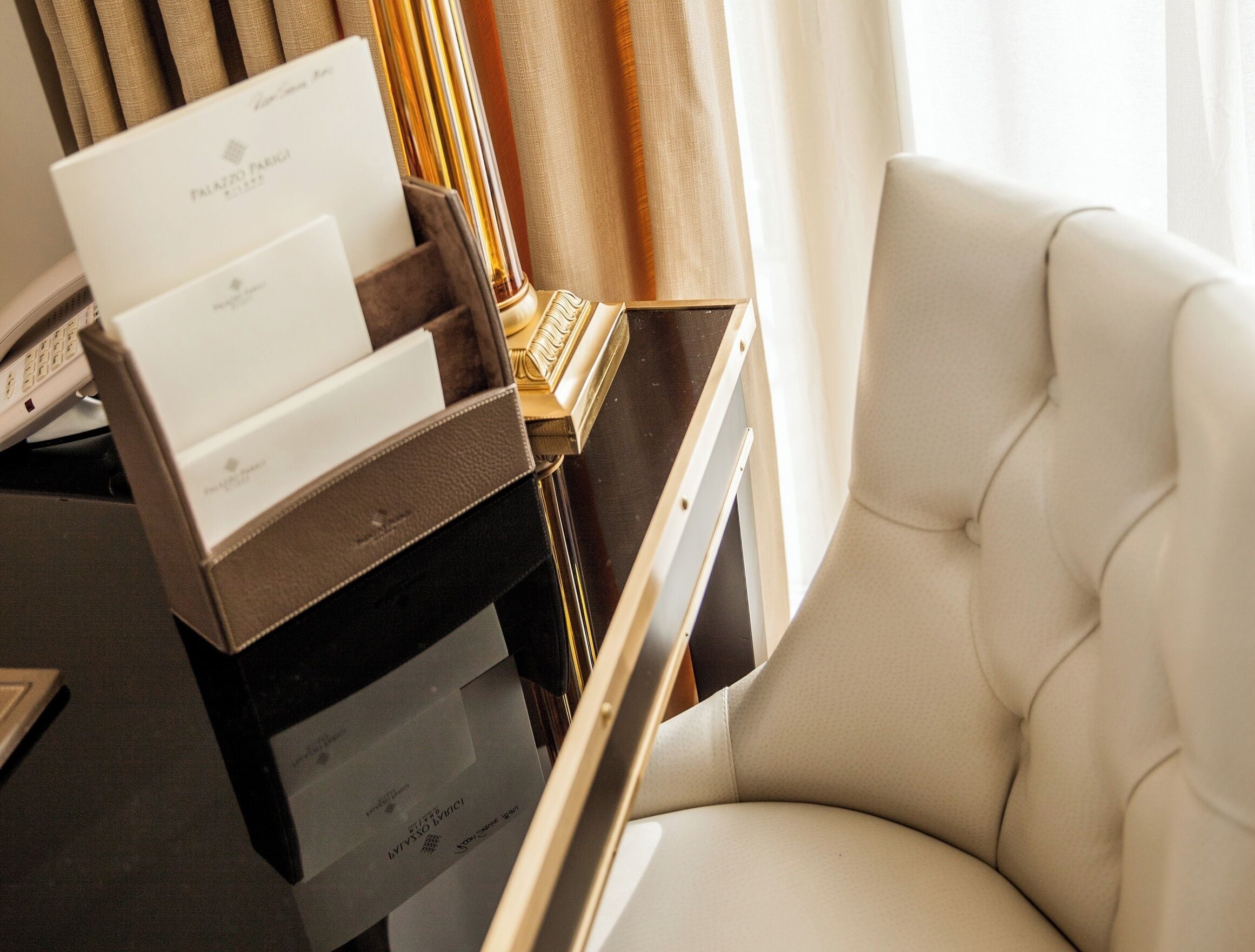 Quarto de hotel com uma poltrona branca macia, uma escrivaninha com um laptop e uma mesinha com apoio para bandejas contendo cartões de visita e um caderno.