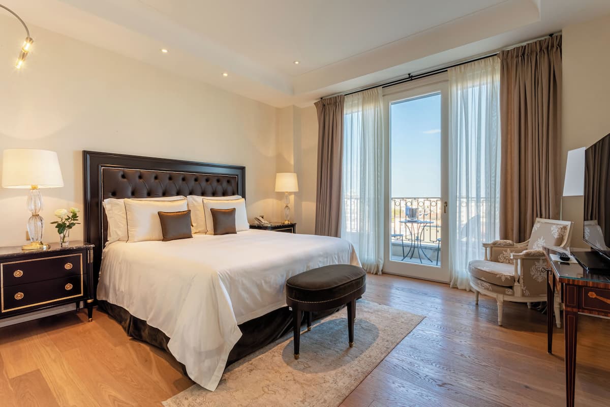 Ein geräumiges Schlafzimmer mit einem großen Bett, einer Sitzecke mit einem Sessel, einem Schreibtisch und einem Balkon mit Vorhängen.