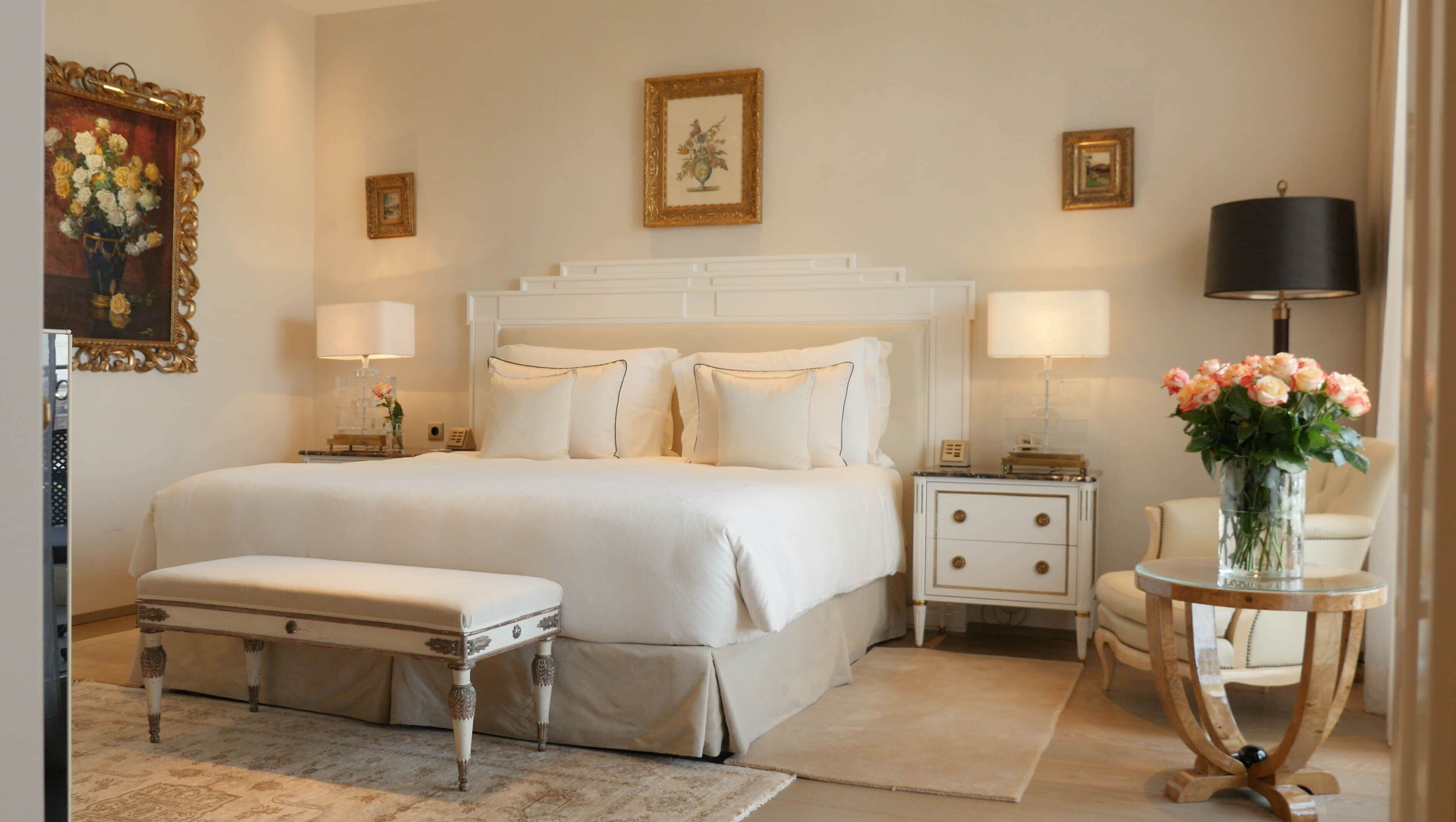 Un dormitorio con una cama grande, mesitas de noche, una silla y un jarrón con flores.