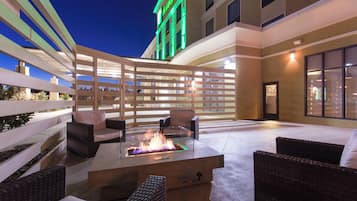 An outdoor patio area with wicker furniture, a glass-top fire feature, and a building with a green-lit sign in the background.