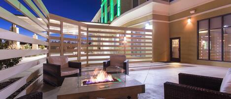 An outdoor patio area with wicker furniture, a glass-top fire feature, and a building with a green-lit sign in the background.