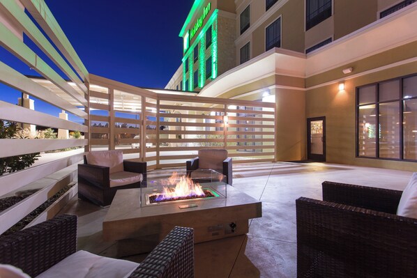 An outdoor patio area with wicker furniture, a glass-top fire feature, and a building with a green-lit sign in the background. - Holiday Inn Texarkana Arkansas Conv Ctr by IHG (Texarkana)