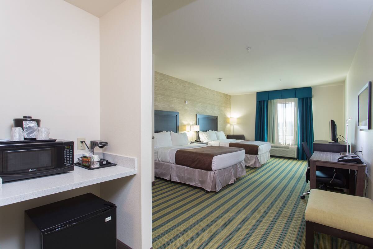 A hotel room with a kitchenette, two beds, a desk, and a window with curtains.