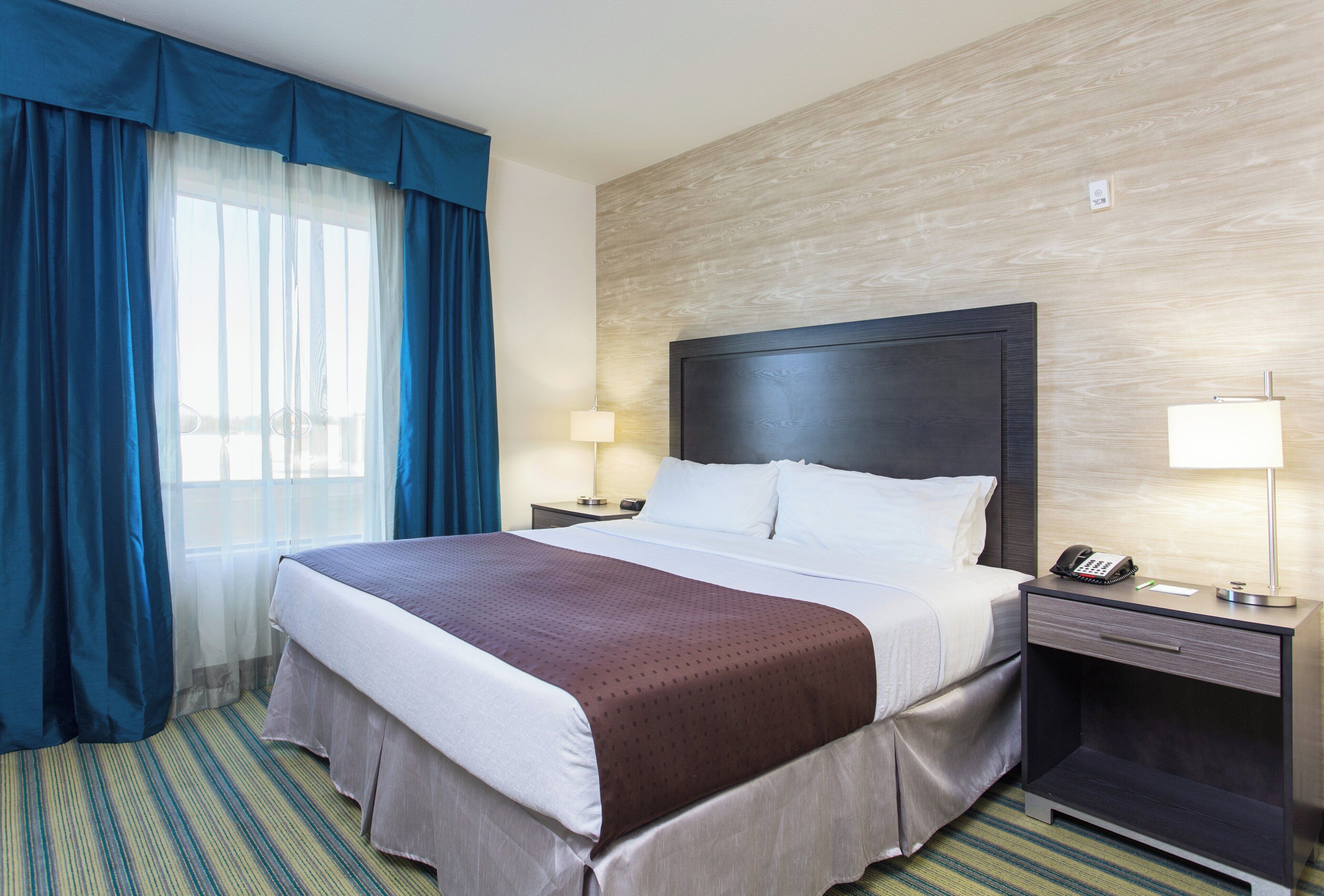Habitación de hotel con una cama grande, una mesita de noche con teléfono, una lámpara y una ventana con cortinas azules.