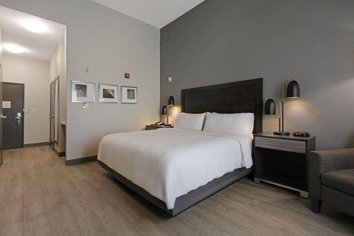 A modern hotel room with a large bed, bedside lamps, a nightstand, and a small armchair.