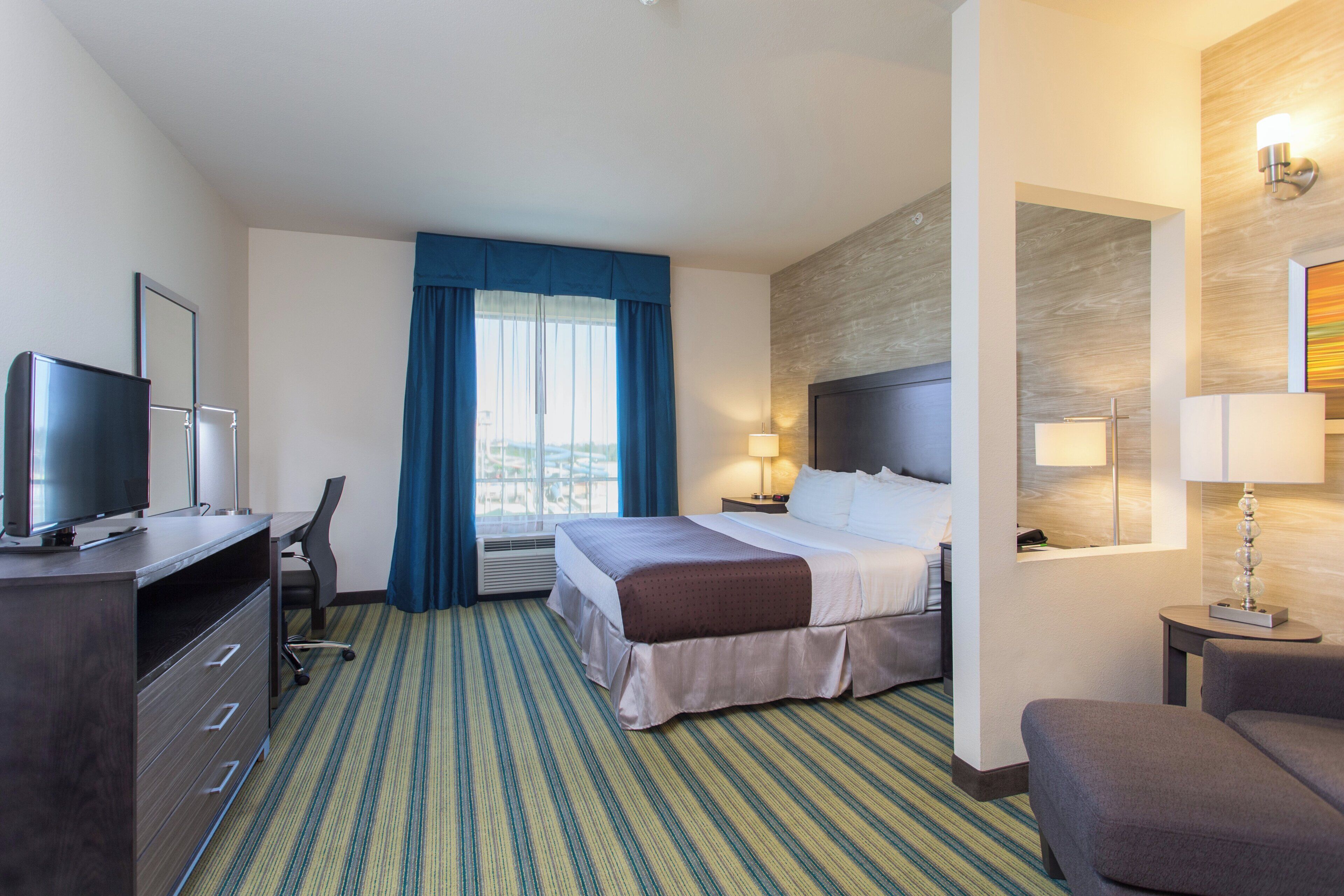 A hotel room with a bed, a desk with a computer, a chair, a TV, and a window with curtains.