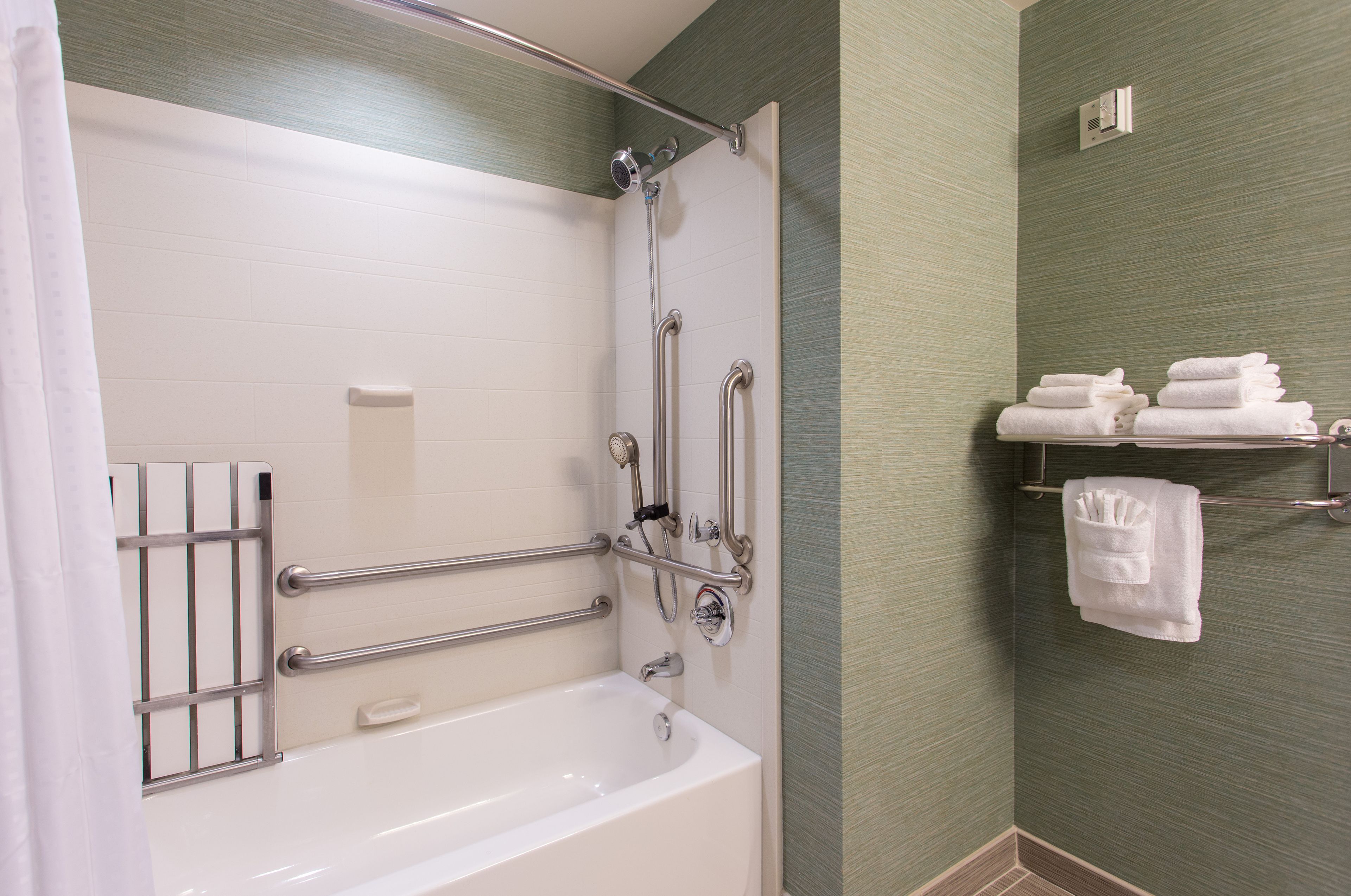 A bathroom with a white bathtub, grab bars, a towel rack with towels, and a shower head.
