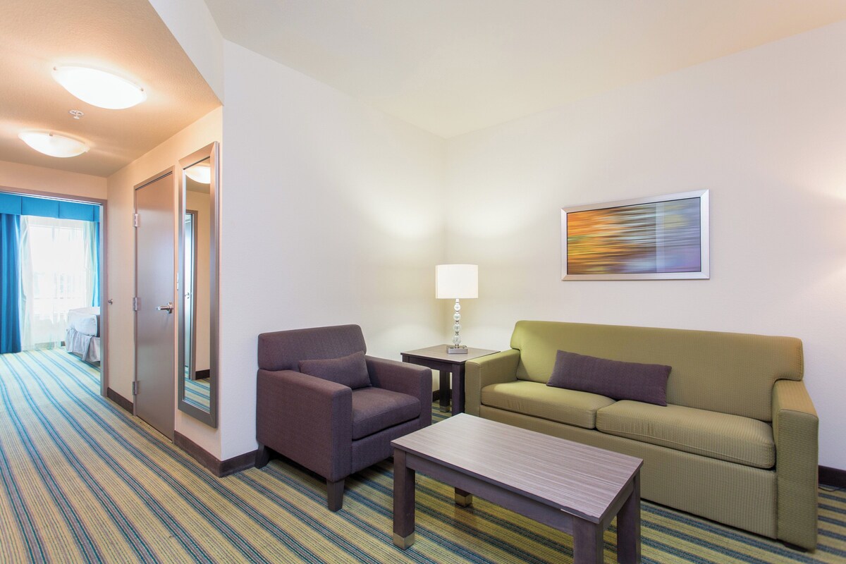 Ein Hotelzimmer mit Sofa, Sessel, kleinem Tisch und einer Lampe. An der Wand hängt ein Gemälde, und eine Tür führt in einen weiteren Raum.