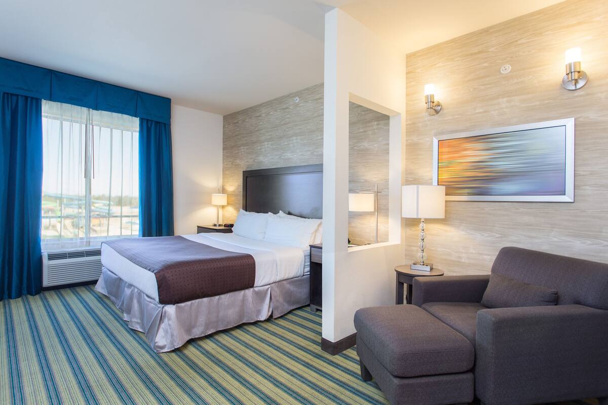 A hotel room with a large bed, two bedside tables with lamps, a gray armchair, a large window with blue curtains, and a painting on the wall.