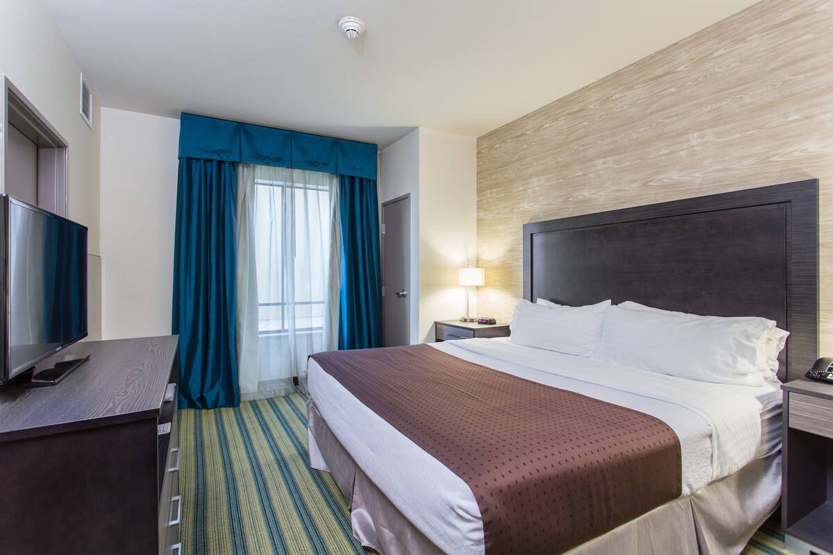 A hotel room with a large bed, a desk with a television, a lamp, and a window with blue curtains.