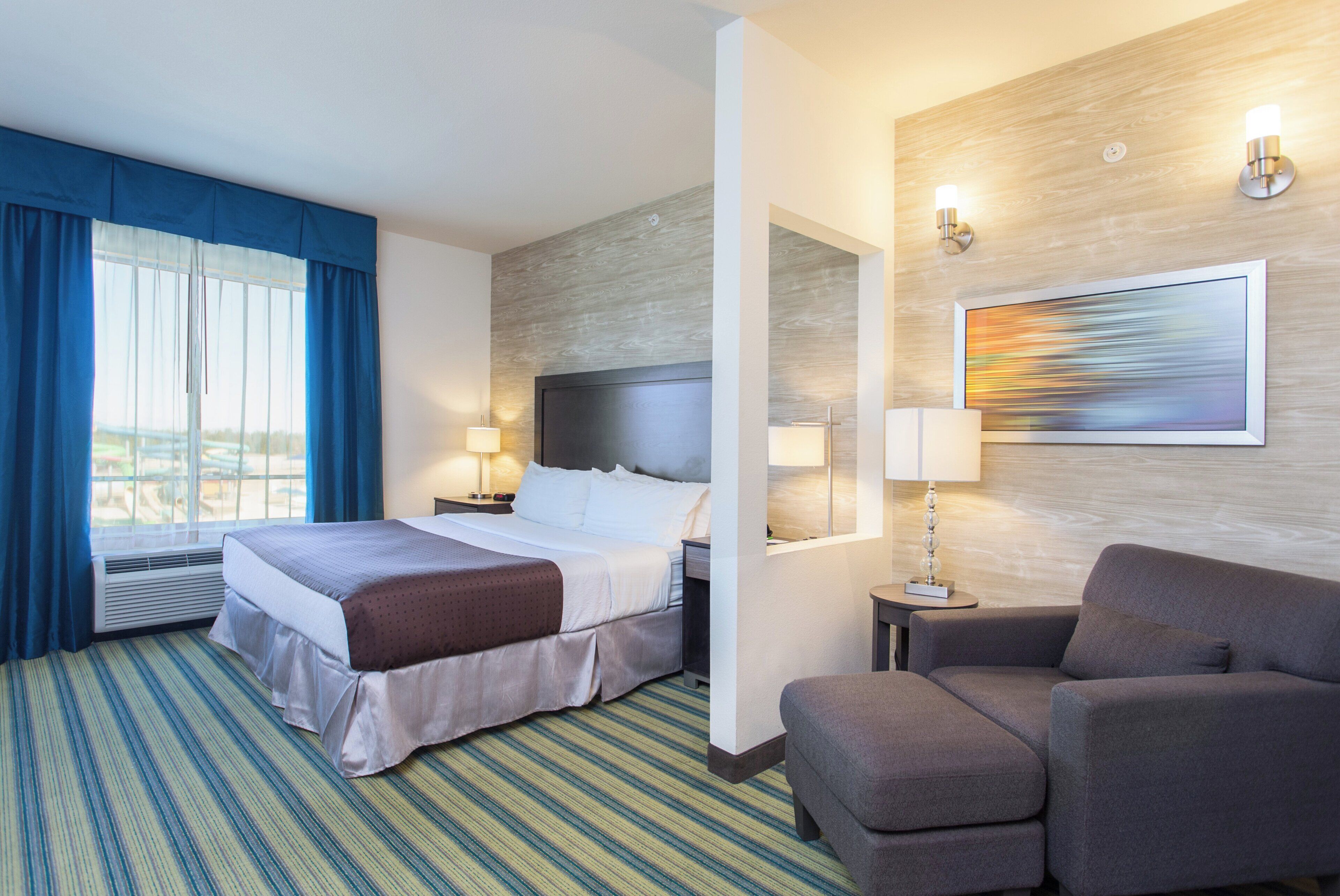 Habitación de hotel con cama, una silla, un escritorio, una lámpara, una ventana con cortinas y un cuadro en la pared.