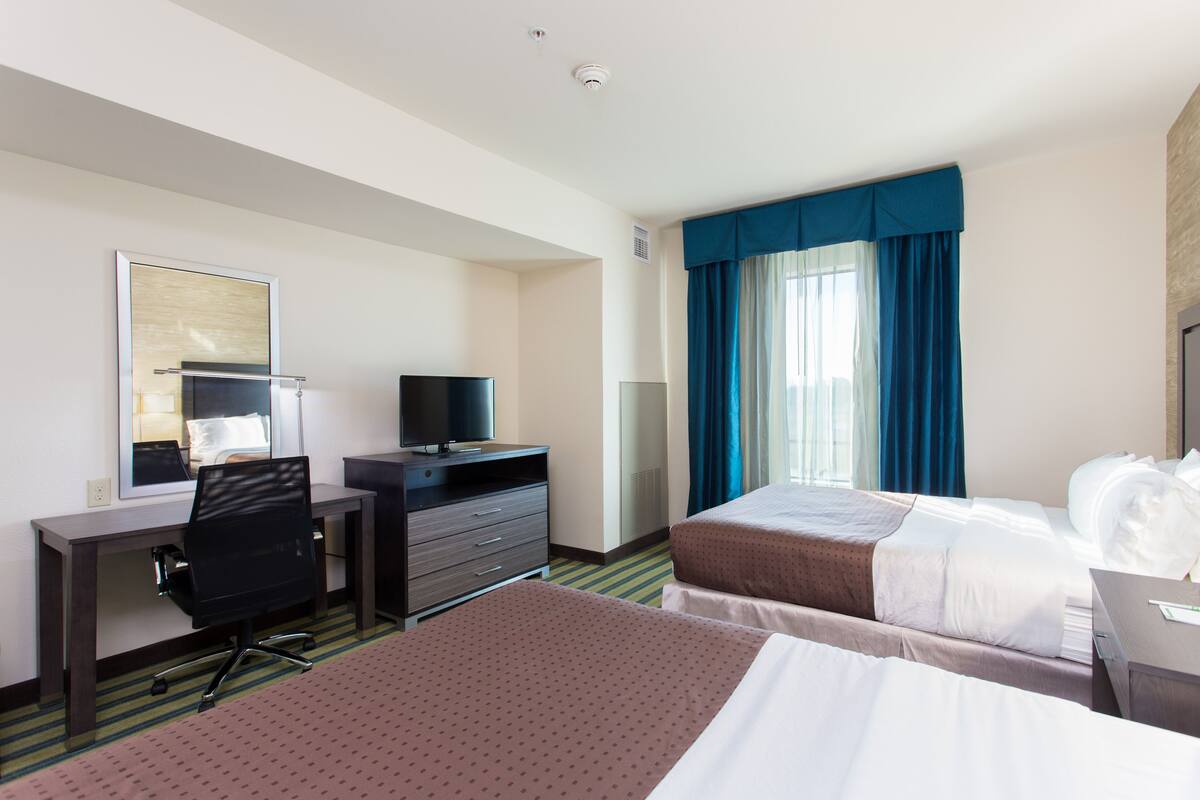 A hotel room with two beds, a desk with a chair, a flat-screen TV, and a large window with curtains.