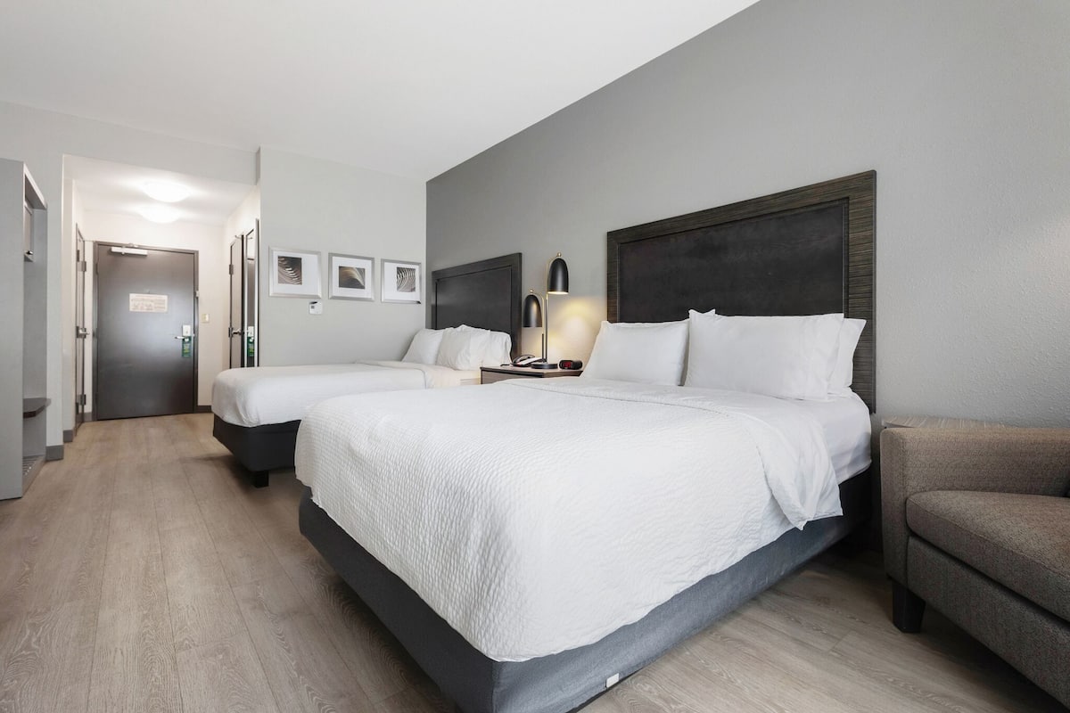 A hotel room with two beds, a sofa, a bedside table with a lamp, and a door leading to a hallway.