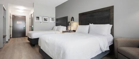 Et hotelværelse med to senge, en sofa, et sengebord med lampe og en dør til en gang.
