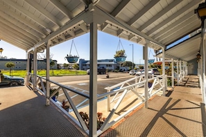 Un espacio exterior cubierto con estructura de metal, macetas colgantes y un estacionamiento al fondo.