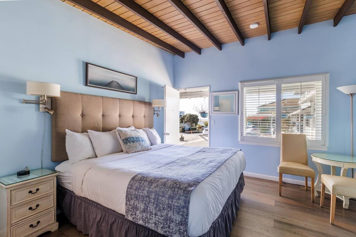 A bedroom with a large bed, a bedside table, a chair, a window with blinds, and a ceiling with wooden beams.
