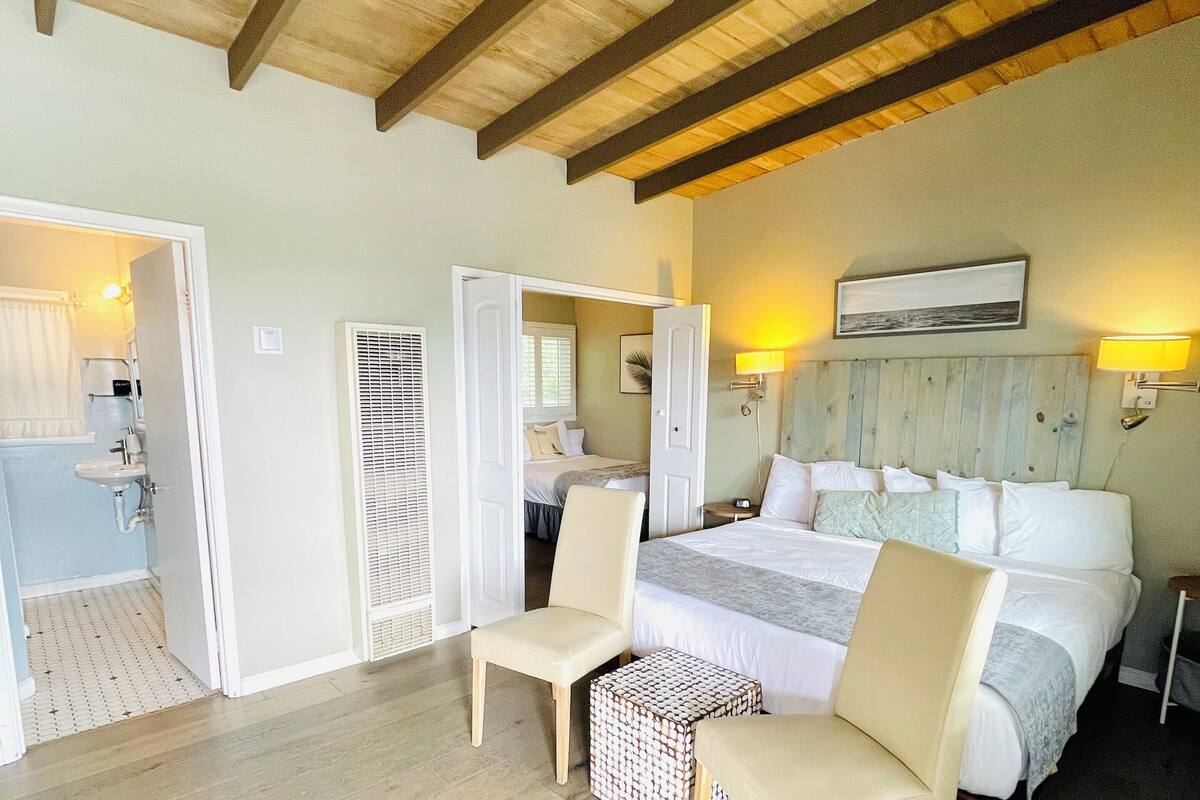 A hotel room with a wooden ceiling, a large bed, two chairs, a small table, and a bathroom visible through an open door.