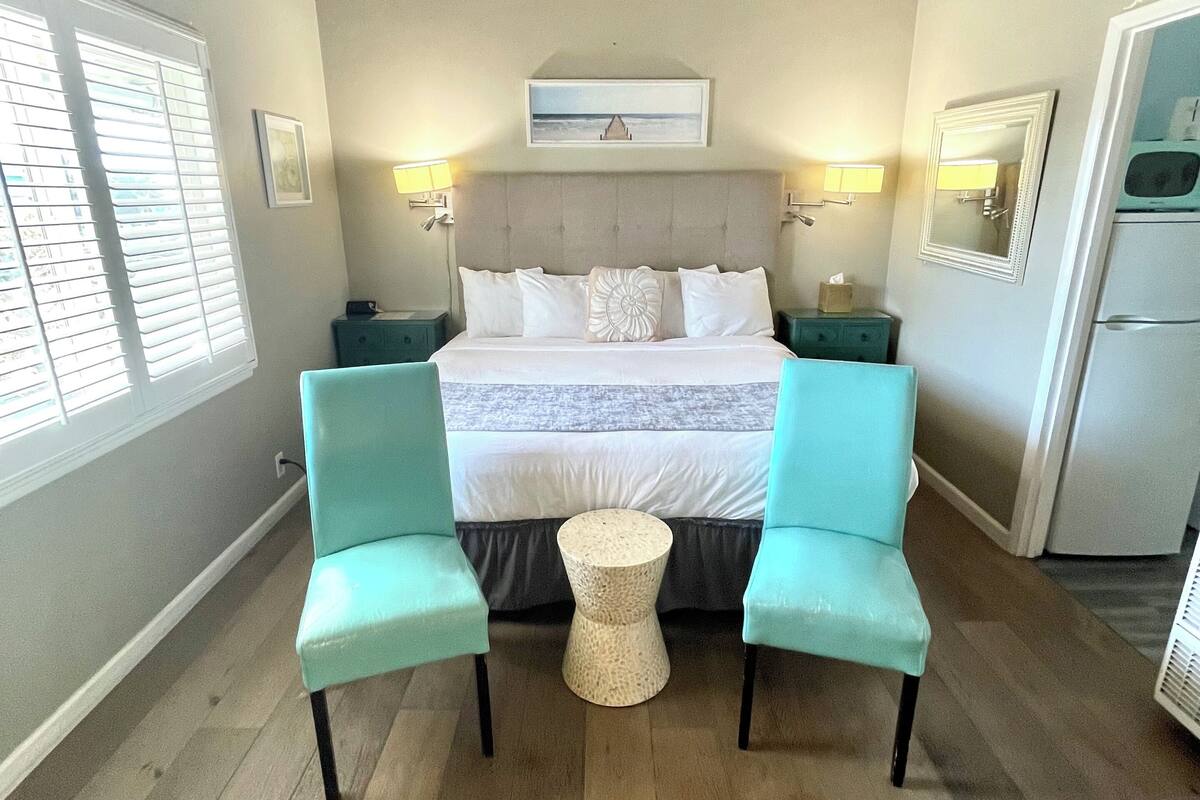 A bedroom with a bed, two chairs, a small round table, a ceiling fan, a mirror, and a window with blinds.