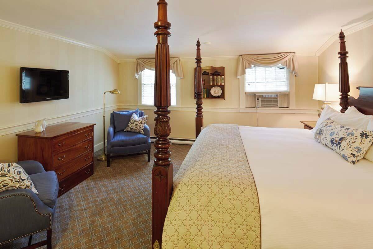 A hotel room with a four-poster bed, a flat-screen TV, a desk with a lamp, a chair, and a bookshelf.