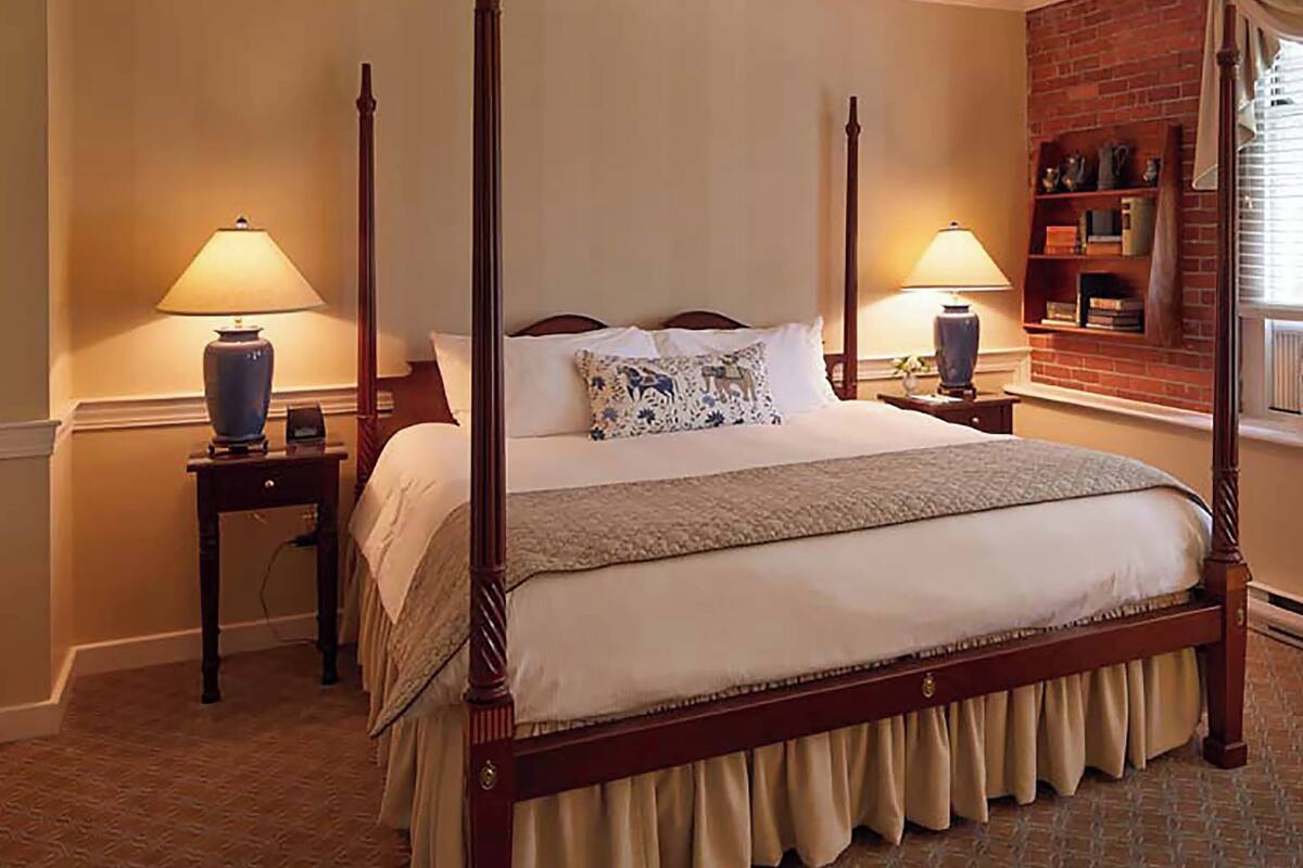 A four-poster bed with a wooden frame, two bedside tables with lamps, a brick wall, and a window with blinds.