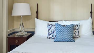 Una cama bien tendida con sábanas blancas y dos almohadas estampadas, una mesita de noche con lámpara y un cabecero de madera.