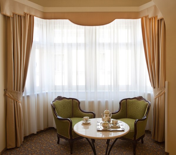 A room with two green upholstered chairs, a round table with a teapot and cups, and a large window with sheer curtains. - Barokk Hotel Promenád Gyor (Gyor)