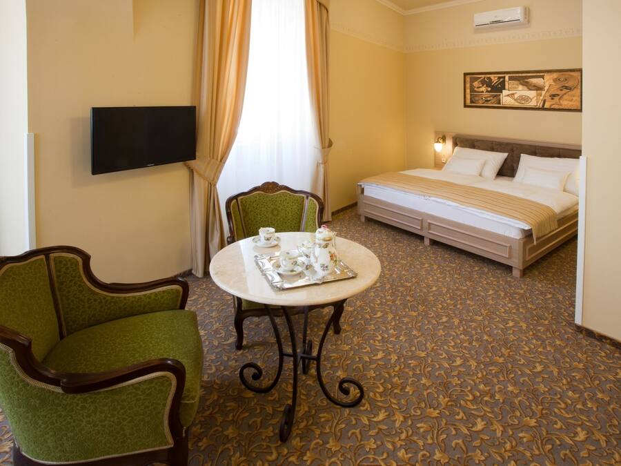 A hotel room with a bed, a TV, a green armchair, a table with teacups, and a patterned carpet.