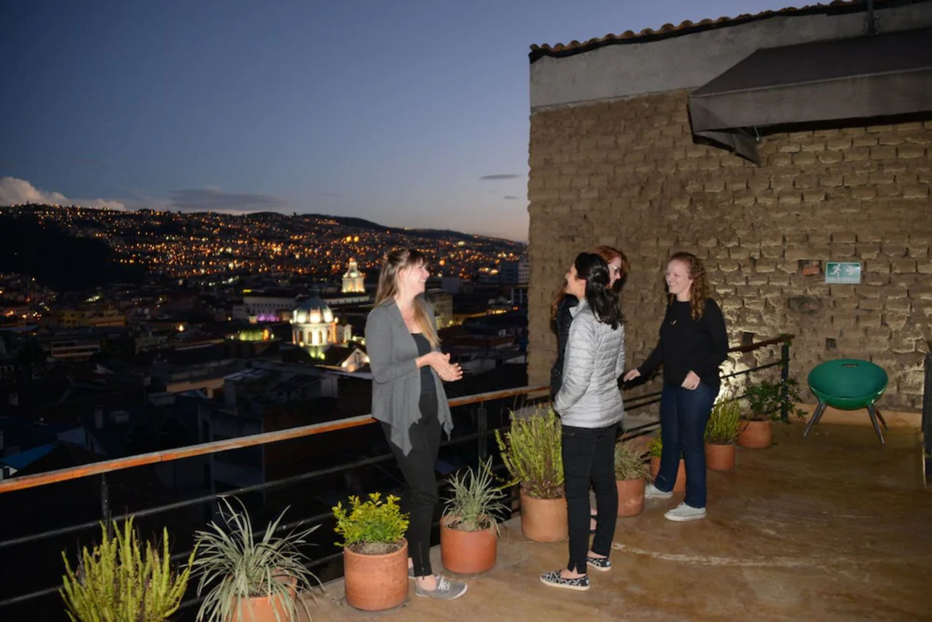 Um grupo de pessoas num terraço com plantas em vasos e vista para a cidade ao entardecer.