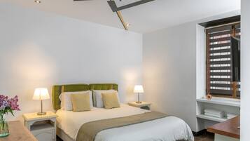 Quarto bem arrumado com uma cama, duas mesas de cabeceira, um criado-mudo, uma janela com persianas e um pequeno espaço de escritório.