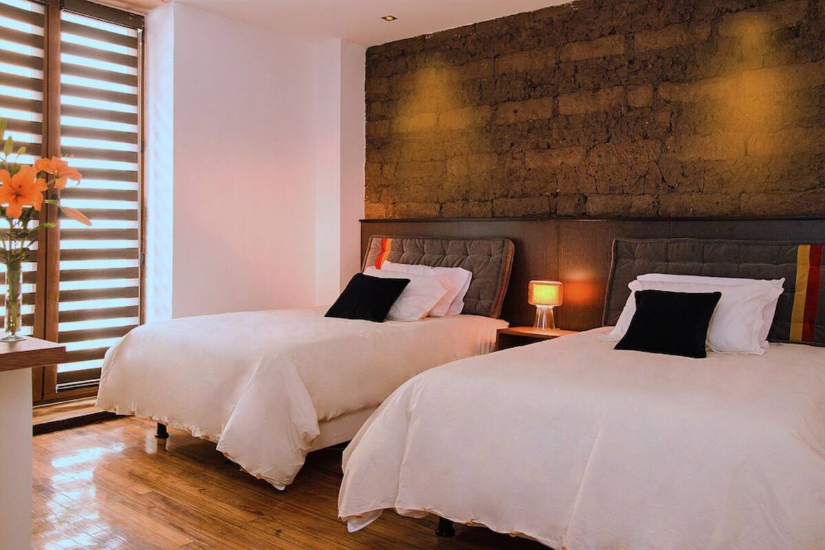A hotel room with two beds, wooden flooring, a brick accent wall, and a bedside table with a lamp.