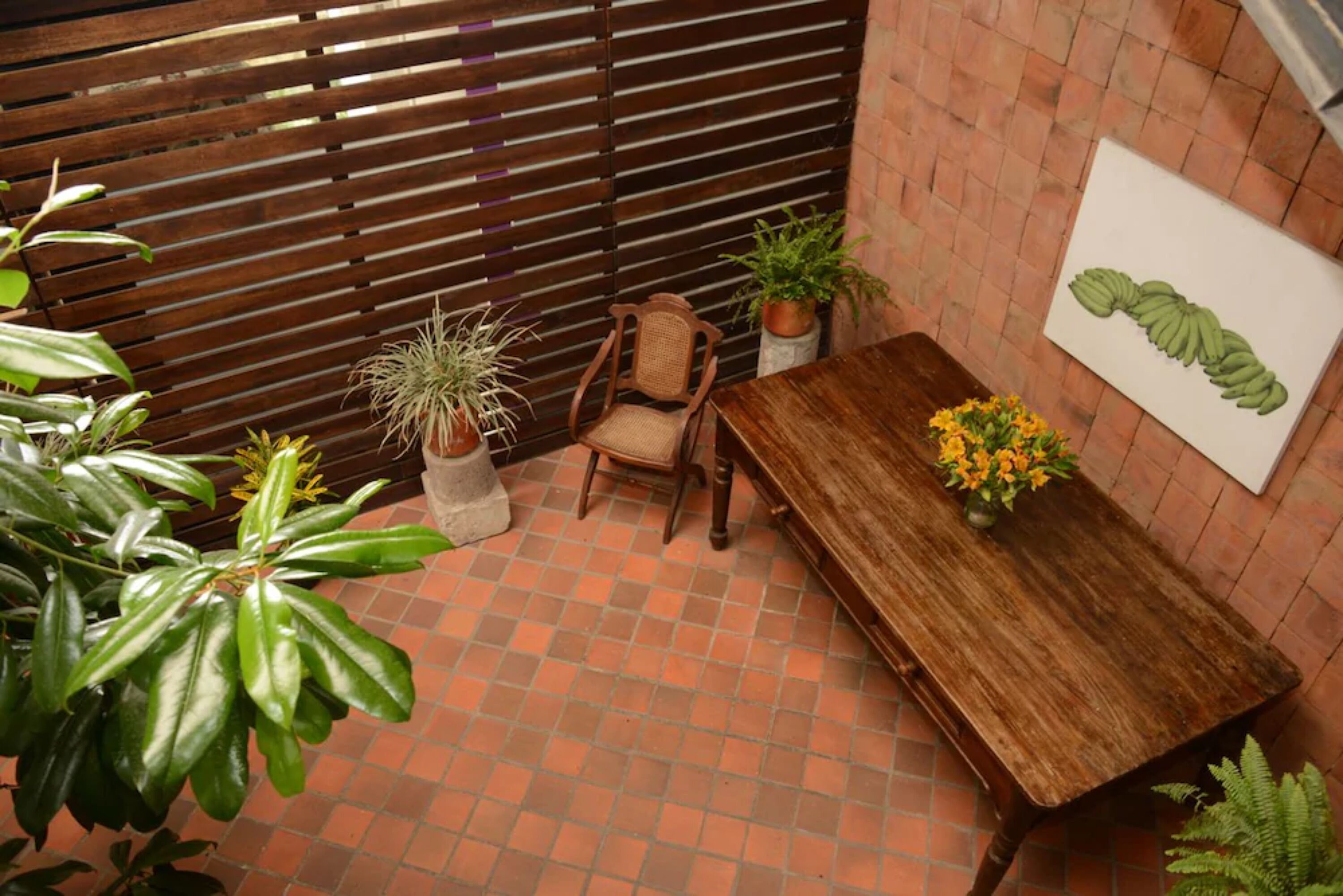 Una mesa de madera con un jarrón de flores amarillas, una silla y plantas en macetas, en una habitación con suelo de baldosas y pared de listones de madera.