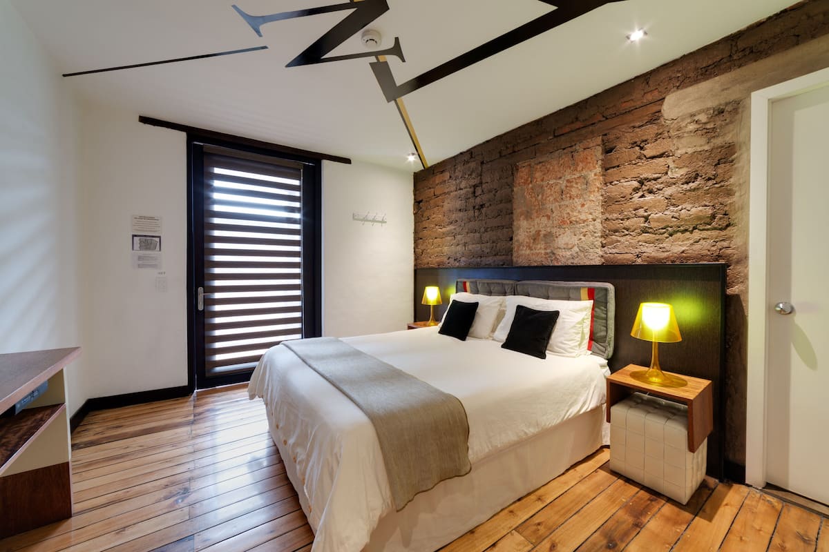 A modern bedroom with a large bed, wooden flooring, a brick accent wall, and a ceiling with exposed beams.