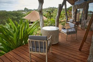 A wooden deck with a round table and two chairs, surrounded by tropical plants and overlooking a thatched-roof building. - Hotel Hare Uta (Hanga Roa)