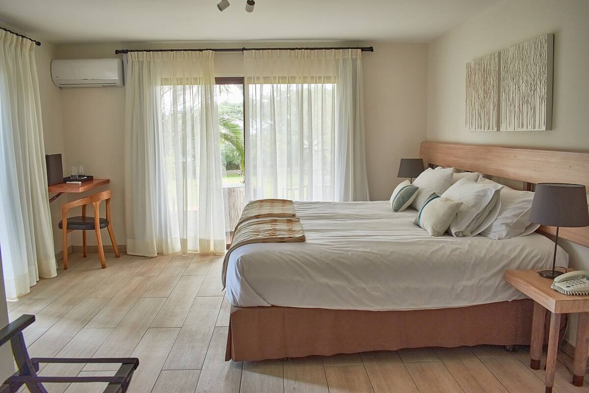 A hotel room with a large bed, a desk, and a chair. There is an air conditioning unit on the wall and a window with curtains.