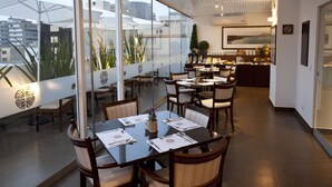 Un restaurant doté d’un mur en verre offrant une vue sur la ville, de tables en bois avec des serviettes blanches et d’un plafond au design moderne.