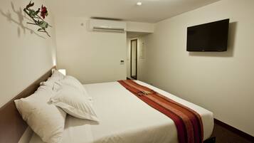 A hotel room with a bed, pillows, a blanket, an air conditioning unit, a flat-screen TV, and a door leading to another room.