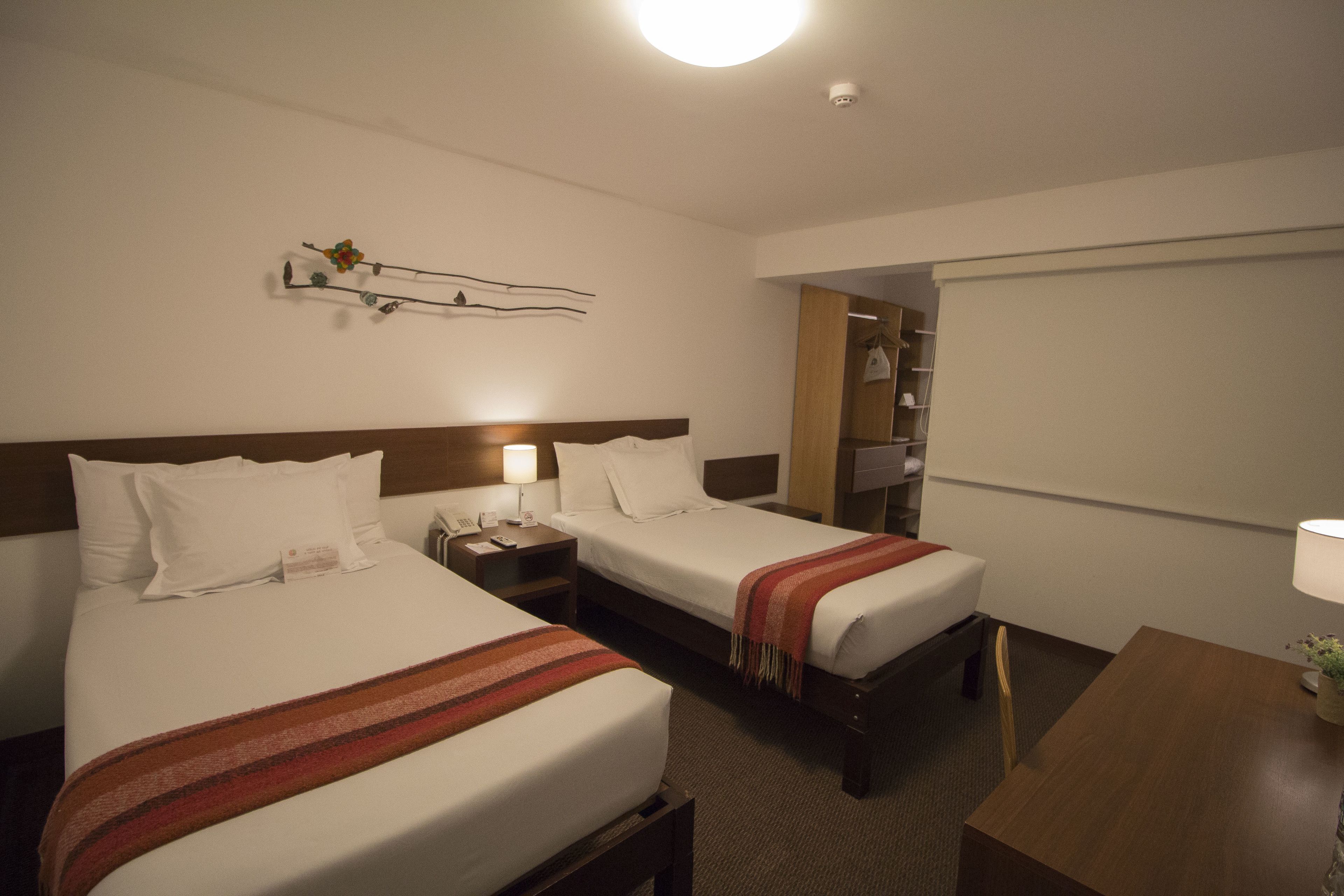 Quarto de hotel com duas camas, uma escrivaninha, uma cadeira, mesa de cabeceira com luminária, prateleira com objetos decorativos e um espelho.