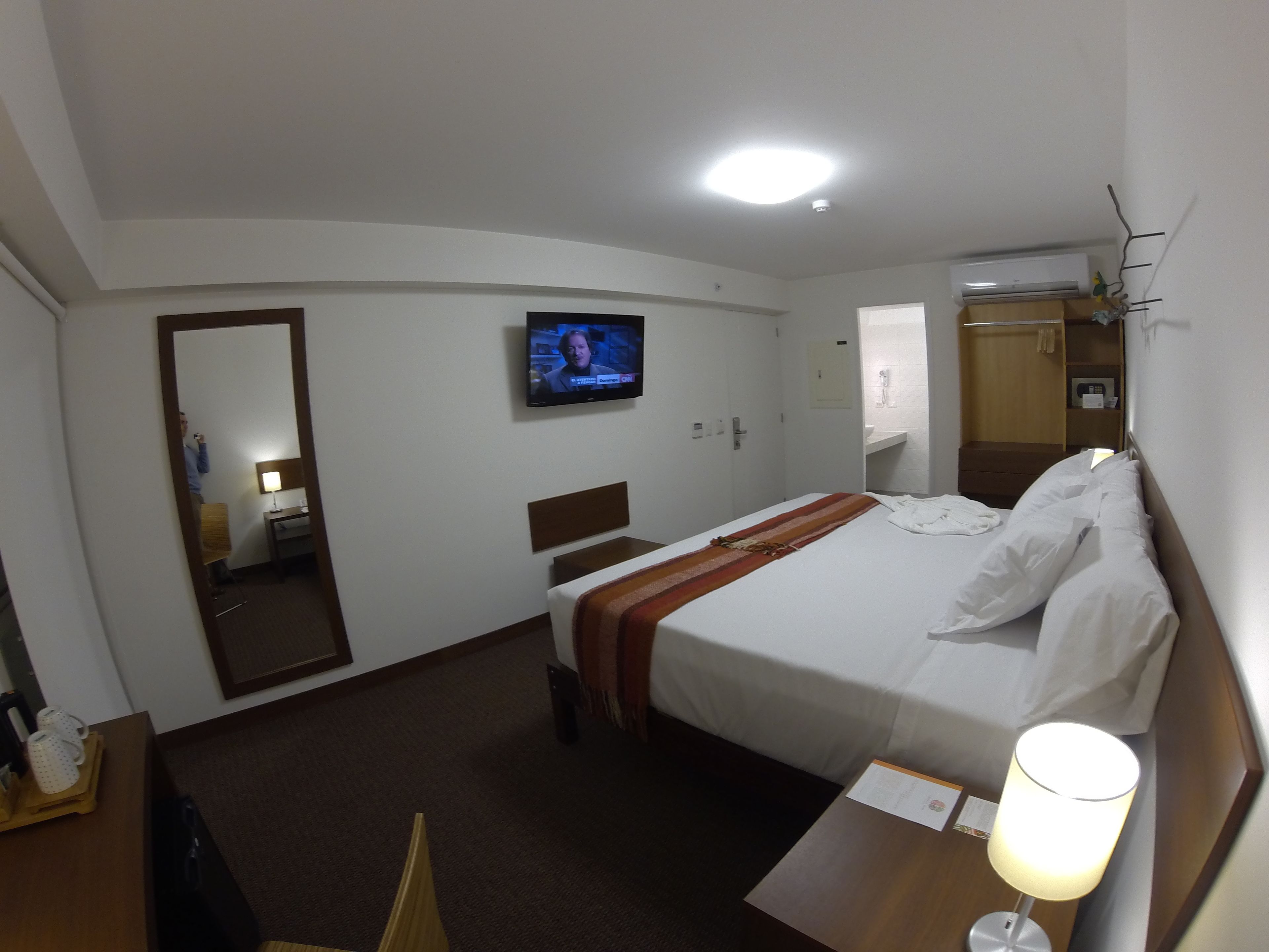 A hotel room with a bed, a TV, a desk, a mirror, and a bathroom visible through an open door.