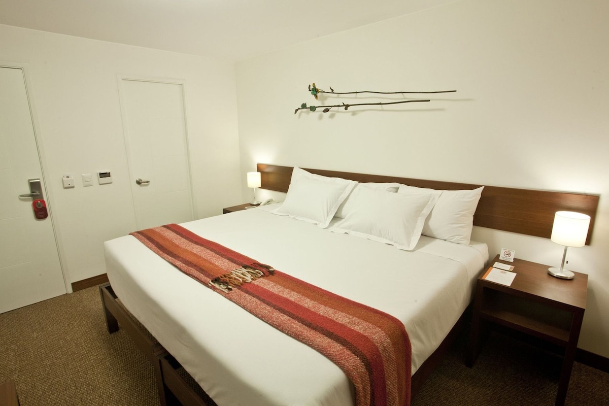 Habitación de hotel con cama, mesita de noche con lámpara, reloj en la pared y puerta con cerradura.