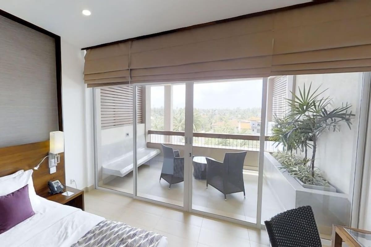 Habitación de hotel con cama, mesita de noche con lámpara, y un amplio ventanal corredero que da a un balcón con silla y una pequeña mesa.