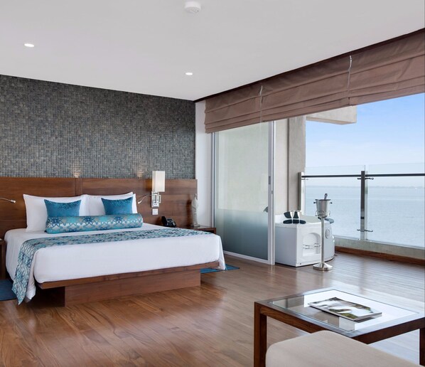 A modern hotel room with a large bed, a bedside lamp, a phone, and a view of the sea through a sliding glass door.