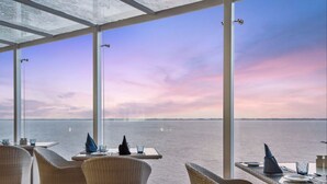 Un restaurant doté de meubles en osier et de grandes fenêtres donnant sur un plan d’eau.