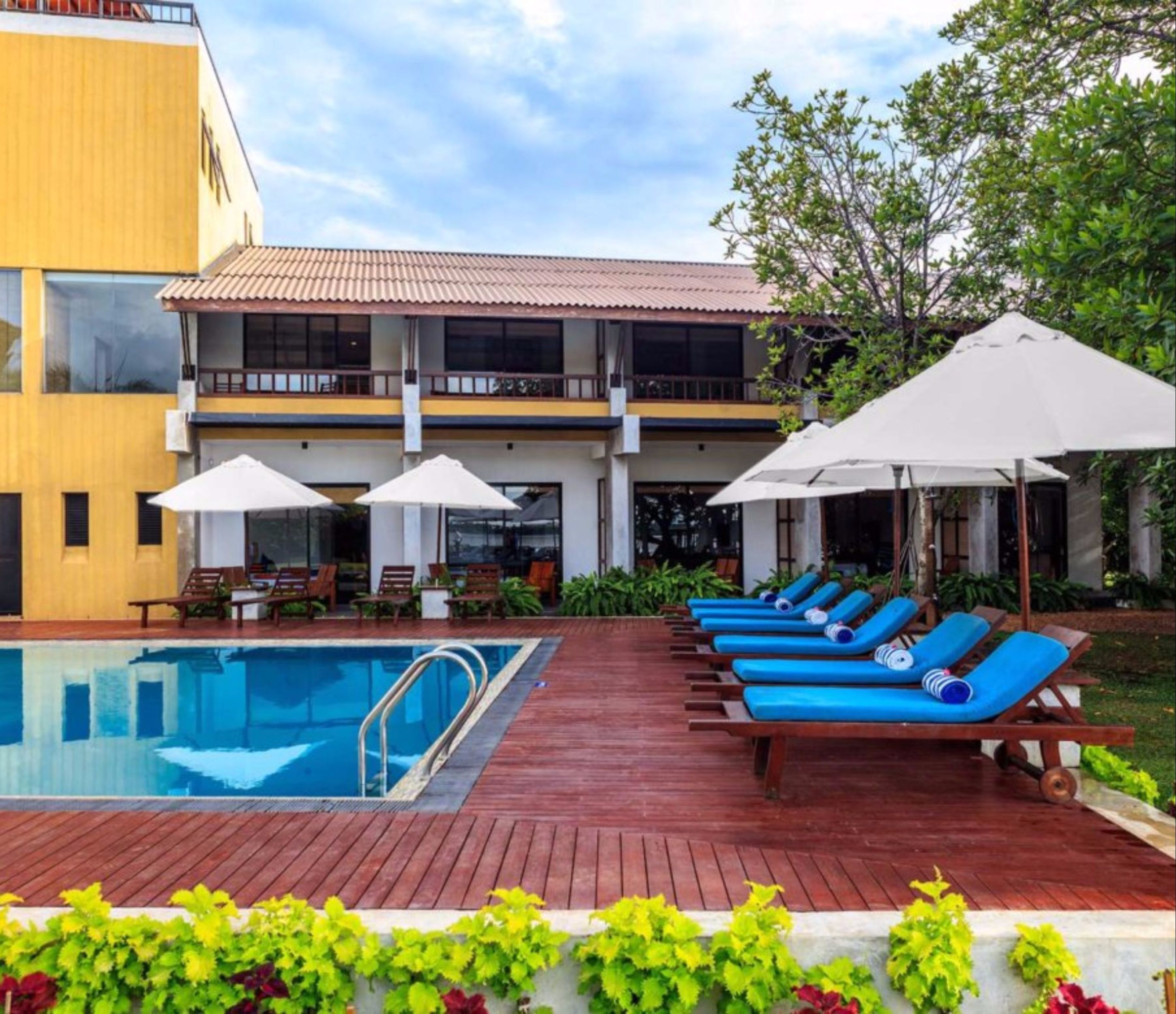 A pool area with a wooden deck, blue lounge chairs, and umbrellas.