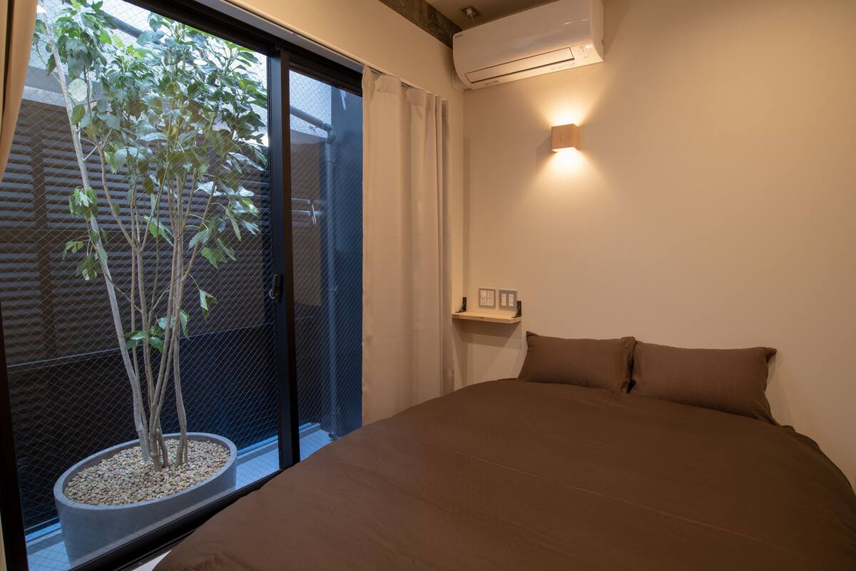 A bedroom with a large bed, a potted plant, a window with curtains, and a wall-mounted air conditioner.